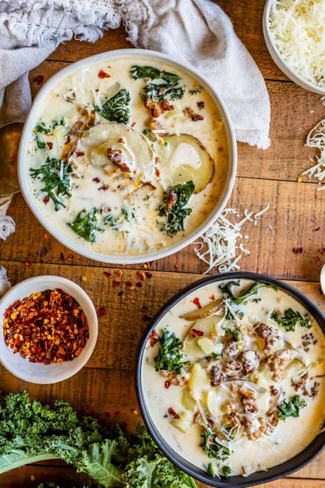 Zuppa Toscana Copycat (Sausage, Potato, and Kale Soup) From The Food Charlatan. Zuppa Toscana is a rich and creamy, soul-satisfying soup made with Italian sausage, potatoes, and hearty kale. It’s a copycat recipe of the famous soup at Olive Garden. I could eat this stuff for days! #zuppatoscana #soup #easy #recipe #olivegarden #onepot #zuppa #toscana #copycat #copy #sausage #potatoes #creamy #cream #halloween #fallsoup #fall #hearty #kale #italiansausage #betterthan sausage kale soup in a white bowl and black bowl on a wooden board with parmesan