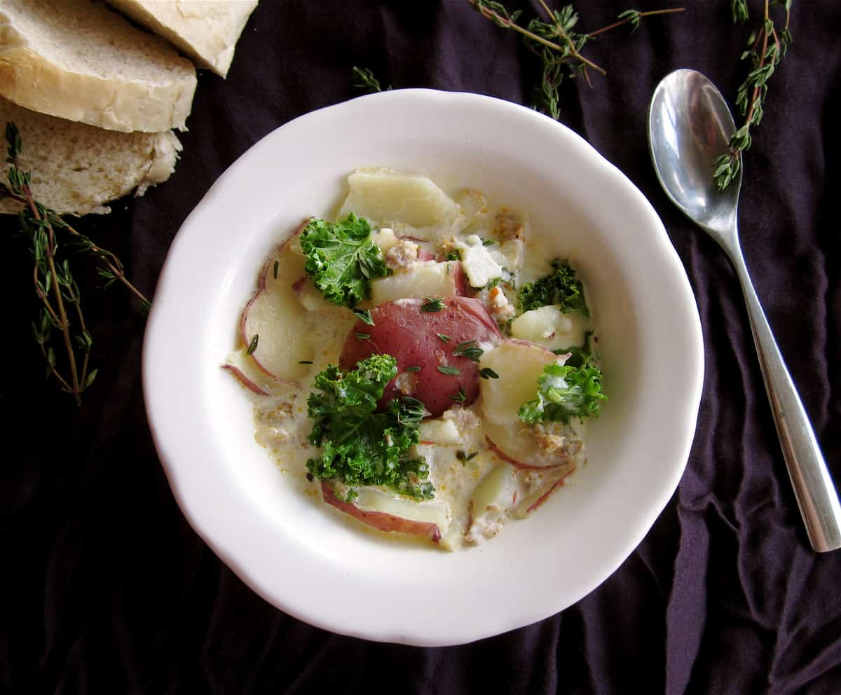 Zuppa Toscana Copycat (Sausage, Potato, and Kale Soup) From The Food Charlatan. Zuppa Toscana is a rich and creamy, soul-satisfying soup made with Italian sausage, potatoes, and hearty kale. It’s a copycat recipe of the famous soup at Olive Garden. I could eat this stuff for days! #zuppatoscana #soup #easy #recipe #olivegarden #onepot #zuppa #toscana #copycat #copy #sausage #potatoes #creamy #cream #halloween #fallsoup #fall #hearty #kale #italiansausage #betterthan Sausage Potato & Kale Soup in a white bowl with spoon and purple napkin
