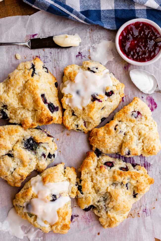 a circle of scones with glaze drizzled over some, butter and jam nearby.
