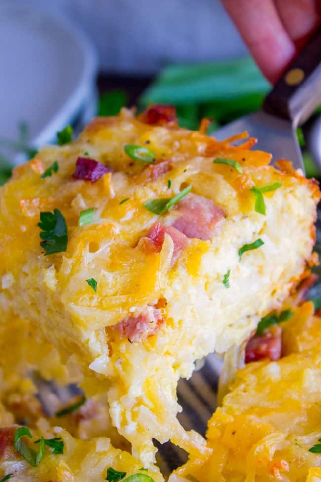 Cheesy Overnight Hashbrown Breakfast Casserole from The Food Charlatan. This Cheesy Hashbrown Breakfast Casserole is everything you need on Christmas morning! Hashbrowns are baked til crispy, then topped with eggs, cheese, and black forest ham. It's an overnight recipe to boot! Make ahead breakfasts ftw. #makeahead #cheese #breakfast #casserole #breakfastcasserole #easy #recipe #ham #blackforestham #Christmas #party #holildays #withhashbrowns #withpotatoes #overnight #makeahead #foracrowd #best breakfast casserole with ham close-up of a piece lifted by a spatula from the pan, connected to casserole by melted cheese.