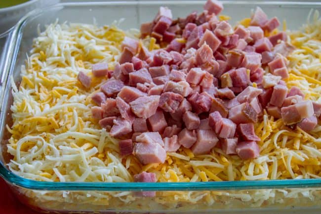 Cheesy Overnight Hashbrown Breakfast Casserole from The Food Charlatan. This Cheesy Hashbrown Breakfast Casserole is everything you need on Christmas morning! Hashbrowns are baked til crispy, then topped with eggs, cheese, and black forest ham. It's an overnight recipe to boot! Make ahead breakfasts ftw. #makeahead #cheese #breakfast #casserole #breakfastcasserole #easy #recipe #ham #blackforestham #Christmas #party #holildays #withhashbrowns #withpotatoes #overnight #makeahead #foracrowd #best christmas casserole in a dish, uncooked, with ham piled over crispy hash browns and shredded cheese.