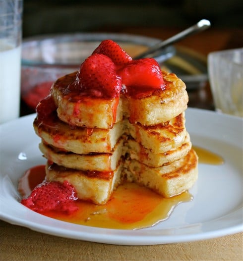 stack of pancakes with strawberries and maple syrup on a white plate.