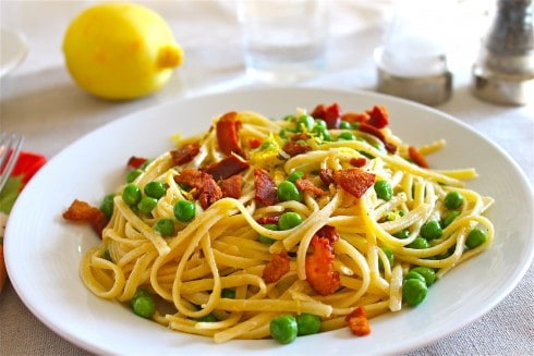 Plate of lemon pasta with bacon and green peas on white tablecloth with lemon in background.