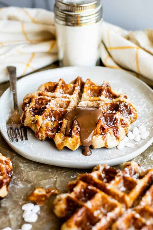 Liege Waffles with Pearl Sugar from The Food Charlatan. Liege waffles are made with a brioche-like yeast dough and are studded with pearl sugar. Pearl sugar is basically a small chunk of sugar that holds its shape as the waffles cooks, leaving you with gloriously crunchy bites of sugar. You can buy these on the street in Belgium and Norway. They make regular waffles look like amateurs. #liege #waffle #pearlsugar #belgium #syrup #maple #easy #recipe #breakfast #brunch #yeast #sugarwaffle liege waffles on a plate with nutella melting on top and pearl and powdered sugars nearby.
