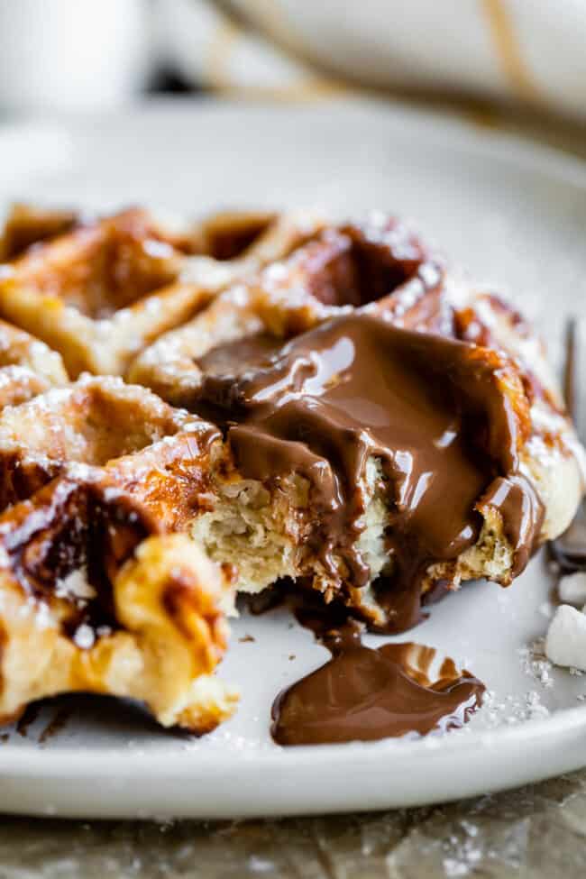 Liege Waffles with Pearl Sugar from The Food Charlatan. Liege waffles are made with a brioche-like yeast dough and are studded with pearl sugar. Pearl sugar is basically a small chunk of sugar that holds its shape as the waffles cooks, leaving you with gloriously crunchy bites of sugar. You can buy these on the street in Belgium and Norway. They make regular waffles look like amateurs. #liege #waffle #pearlsugar #belgium #syrup #maple #easy #recipe #breakfast #brunch #yeast #sugarwaffle a liege waffle on a plate with nutella melting on top and pearl sugar nearby.