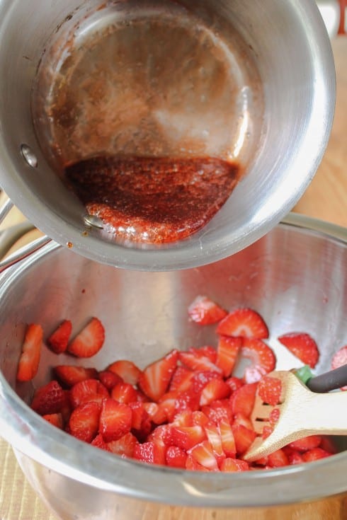Pouring melted jam into a bowl of fresh sliced strawberries.