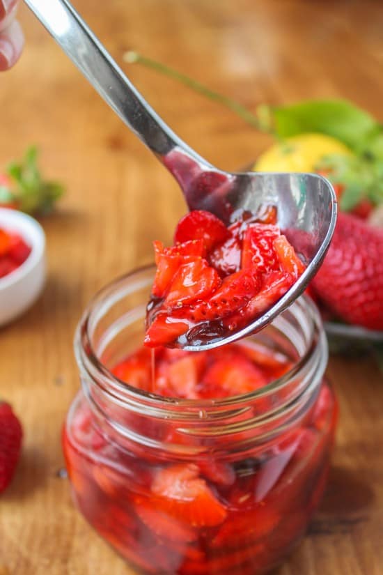 A spoon with strawberry sauce and glass jar of sauce on a cutting board.