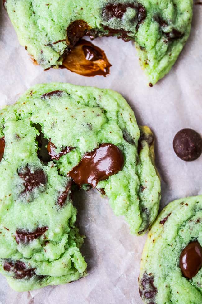 Mint Chocolate Chip Cookies from The Food Charlatan. These Mint Chocolate Chip Cookies are like eating the ice cream in the form of a warm, buttery, gooey cookie. They're crisp on the edges while perfectly tender and chewy in the center. #mint #chocolate #chip #cookies #peppermint #chocolatechip #gooey #chewy #easy #recipe #crisp two green Chocolate Chip Cookies shot from overhead, one broken