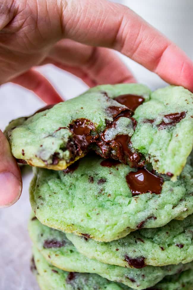 Mint Chocolate Chip Cookies from The Food Charlatan. These Mint Chocolate Chip Cookies are like eating the ice cream in the form of a warm, buttery, gooey cookie. They're crisp on the edges while perfectly tender and chewy in the center. #mint #chocolate #chip #cookies #peppermint #chocolatechip #gooey #chewy #easy #recipe #crisp green chocolate chip cookies stacked with hand holding top one, mint flavored