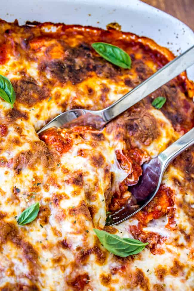 Easy Ravioli Lasagna from The Food Charlatan. This easy ravioli lasagna bake is a hearty no-brainer dinner to make on busy back-to-school nights. You don't even have to cook the ravioli, just throw it in frozen. It's a total crowd-pleaser! And a great make-ahead meal too. #ravioli #lasagna #frozen #quickdinner #ricotta #easy #cheese #mozzarella #marinara #freezermeal #makeahead two metal spoons in a large pan of ravioli lasagna.