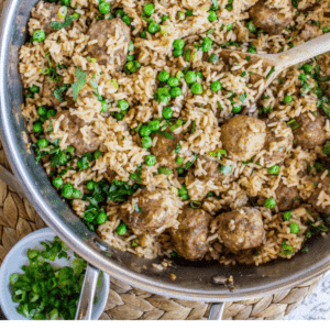 A skillet filled with beef meatballs, rice, and green peas, topped with fresh herbs. A wooden spoon rests inside, and sliced green onions are nearby. Text overlay reads, “Easy One Skillet Beef Meatballs with Rice & Peas.”.