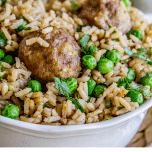 A bowl filled with rice, green peas, and beef meatballs, topped with chopped green onions. Text on the image reads, Easy One Skillet Beef Meatballs with Rice & Peas - The Food Charlatan. Perfect for a quick dinner.