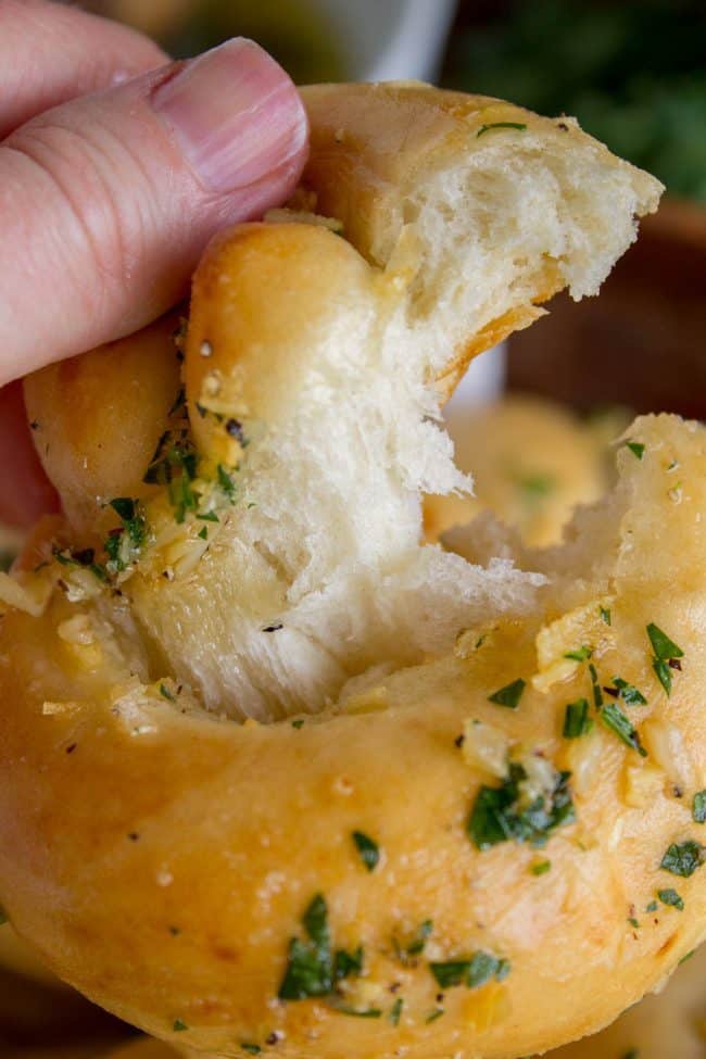 Garlic Knots Recipe from The Food Charlatan. These Garlic Knots are the perfect rolls for your Thanksgiving (or Easter) table! They are chewy and soft, and have TONS of garlicky, buttery flavor! You can't eat just one. (At least I can't. A batch of these rolls is basically a meal for me.) #garlic #bread #knots #rolls #homemade #fromscratch #dinner #holidays #thanksgiving #easter #frozen a hand tearing open a homemade garlic knots with parsley garlic butter.