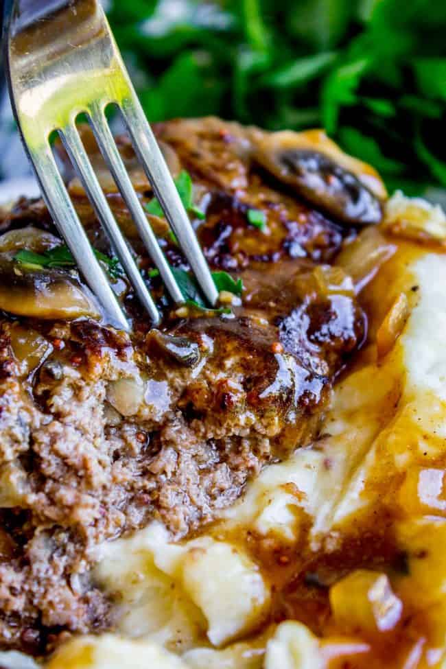 Salisbury Steak with Mushroom Gravy (30 Minute dinner) from The Food Charlatan. Salisbury Steak sounds so fancy but it's SO easy to make. They are basically mini meatloaves fried and served with mushroom gravy. It's the perfect comfort food and only takes 30 minutes to make. You have to serve this with Creamy Mashed Potatoes to get the full effect! #ComfortFood #Easy #30minutes #dinner #oneskillet #beef #groundbeef #mashedpotatoes #gravy #mushrooms #sauce #hamburger #fromscratch salisbury steak with mushroom gravy on a creamy mashed potatoes with a fork lifting a bite.
