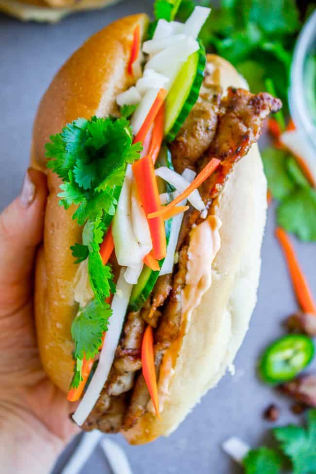 Banh Mi Sandwich from The Food Charlatan. These traditional Vietnamese Banh Mi Sandwiches will blow your mind! A mini soft baguette, smeared with sriracha mayo, topped with thinly sliced marinated pork, pickled veggies, and tons of cilantro. It's heaven! #vietnam #banhmi #sandwich #vietnamese #cilantro #daikon #pickled #carrots #baguette #recipe #pork #howtomake #pickledvegetables #sauce #sriracha #mayonnaise banh mi sandwich held in a hand.