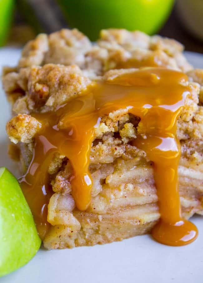 A slice of apple crumble topped with caramel sauce, with layers of baked apples visible, sits on a plate next to a wedge of green apple, resembling delicious apple pie bars.