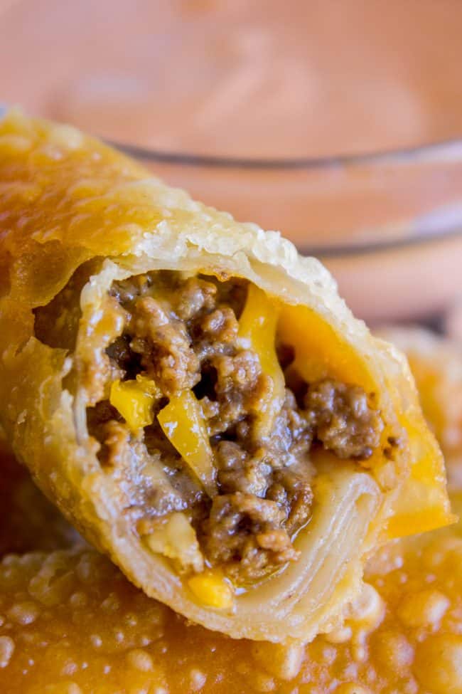 Close-up of crispy cheeseburger egg rolls cut open, revealing a savory filling of ground beef, melted cheese, and yellow bell pepper, with dipping sauce in the background.