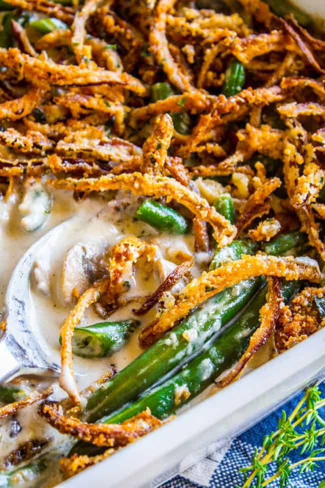 Close-up of green bean casserole with fresh green beans, topped with crispy fried onions and creamy sauce, as a serving spoon scoops out a portion from the baking dish.