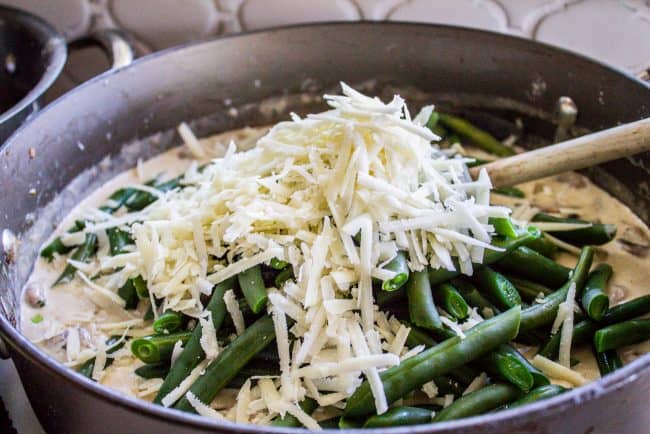 Homemade Green Bean Casserole Recipe from The Food Charlatan. Not your mama's Green Bean Casserole recipe! Let's kick that can of soup out to the curb because the homemade version is LEGIT you guys. Big, bright green beans in a creamy mushroom sauce, topped with crispy fried onions. Thanksgiving doesn't get any better than this! #greenbeans #casserole #frenchs #fromscratch #homemade #thanksgiving #sidedish #easy #fresh #recipe #classic #best #withcheese #foracrowd #makeahead #simple #classic #creamy adding parmesan cheese and blanched green beans to creamy mushroom sauce in a pan.