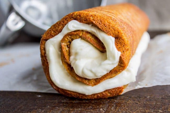 Pumpkin Roll Recipe with Lots of Cream Cheese Frosting from The Food Charlatan. This easy Pumpkin Roll recipe is so impressive and delicious! It's a classic Thanksgiving dessert that you definitely need to make this year. I used LOTS of cream cheese frosting so that it's super moist! #pumpkin #roll #creamcheese #icing #frosting #thanksgiving #christmas #fall #fallbaking #recipe #withcreamcheesefilling #easy #withnutmeg #Libbys #best #homemade #classic #moist #gift #perfect #howtomake #cinnamon rolling a pumpkin roll with cream cheese frosting inside on a piece of parchment paper.