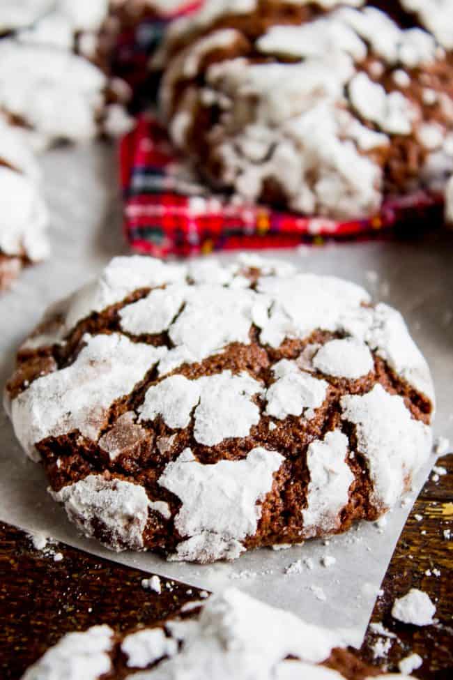 Chocolate Crinkle Cookies (Ultra Fudgy) from The Food Charlatan. It's not even Christmas with Chocolate Crinkle Cookies! They are just a classic. Chocolate Christmas cookies are a must on your cookie plate; they always seem to disappear first. This recipe is ultra fudgy and decadent because we're using cocoa AND baking chocolate! #easy #recipe #classic #chocolate #fudge #fudgy #powderedsugar #crinkle #best #christmas #chewy #brownie #hersheys #withcocoapowder #fromscratch chocolate crinkle cookie on parchment paper.