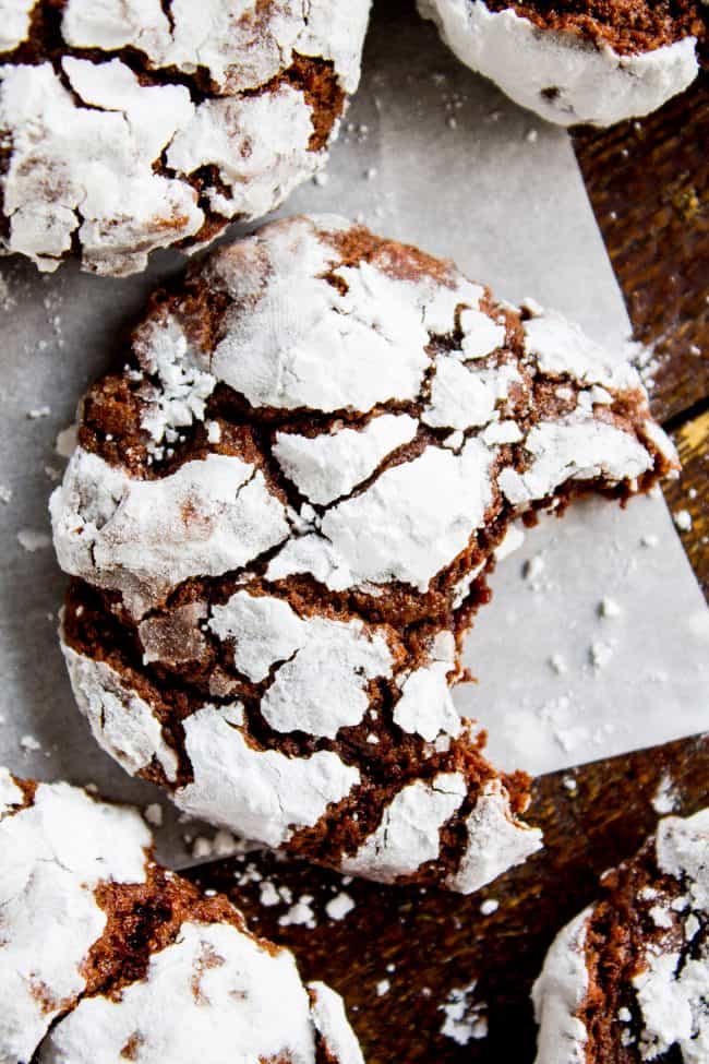 Chocolate Crinkle Cookies (Ultra Fudgy) from The Food Charlatan. It's not even Christmas with Chocolate Crinkle Cookies! They are just a classic. Chocolate Christmas cookies are a must on your cookie plate; they always seem to disappear first. This recipe is ultra fudgy and decadent because we're using cocoa AND baking chocolate! #easy #recipe #classic #chocolate #fudge #fudgy #powderedsugar #crinkle #best #christmas #chewy #brownie #hersheys #withcocoapowder #fromscratch chocolate crinkle cookies on a sheet of parchment paper, one with a bite taken out of it.