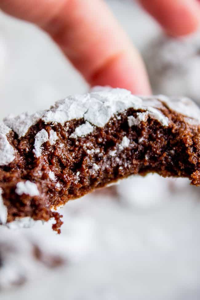 Chocolate Crinkle Cookies (Ultra Fudgy) from The Food Charlatan. It's not even Christmas with Chocolate Crinkle Cookies! They are just a classic. Chocolate Christmas cookies are a must on your cookie plate; they always seem to disappear first. This recipe is ultra fudgy and decadent because we're using cocoa AND baking chocolate! #easy #recipe #classic #chocolate #fudge #fudgy #powderedsugar #crinkle #best #christmas #chewy #brownie #hersheys #withcocoapowder #fromscratch chocolate crinkle cookie with a bite taken out of it.