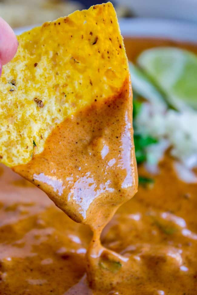 A close-up of a hand holding a tortilla chip dipped in creamy, orange queso dip, with lime wedges and chopped herbs blurred in the background.