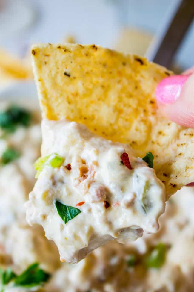 A close-up of a hand holding a tortilla chip dipped in a thick, textured sausage cream cheese dip with chopped green onions and herbs. The creamy dip clings to the chip, and the fingers are polished with pink nail color.