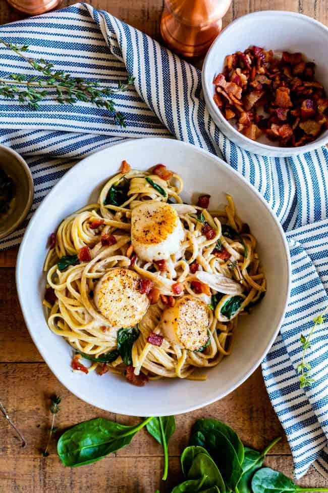 Pan Seared Scallops in Creamy Champagne Sauce from The Food Charlatan. If you have never paired tender, buttery, pan seared scallops with a creamy wine sauce, you are missing out! Toss with some linguine and crispy crumbled bacon, and you have a very special dinner for two. #valentinesday #valentine #dinnerfortwo #romantic #scallops #recipe #easy #pansear #seafood #seared #healthy #pasta #Howtocook #dinner #easy #fried #best #sauteed #creamy #spinach #meal #whattoserve #whitewine #champagne scallops with linguine, bacon, and spinach on a striped towel with a bowl of crumbled bacon.