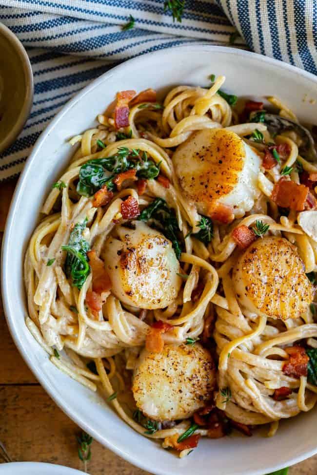 Pan Seared Scallops in Creamy Champagne Sauce from The Food Charlatan. If you have never paired tender, buttery, pan seared scallops with a creamy wine sauce, you are missing out! Toss with some linguine and crispy crumbled bacon, and you have a very special dinner for two. #valentinesday #valentine #dinnerfortwo #romantic #scallops #recipe #easy #pansear #seafood #seared #healthy #pasta #Howtocook #dinner #easy #fried #best #sauteed #creamy #spinach #meal #whattoserve #whitewine #champagne pan seared scallops with creamy wine sauce, bacon, and spinach over linguine in a white bowl.