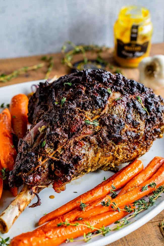 Roast Leg of Lamb with Blackened Honey Dijon Crust from The Food Charlatan. #ad #OhMaille #MyMaille #IDidItMailleWay Roast leg of lamb makes an unforgettable main dish for your Easter celebration! Roast lamb is so delicious, moist, and tender when it is done right. The secret is to turn the oven to 500 degrees to get that blackened honey dijon crust made with Maille® Honey Dijon, then lower it to get the most tender, juicy roast lamb you've ever had. #Walmart #leg #recipes #dinner #easter #lamb whole roast leg of lamb on a platter with carrots and thyme.