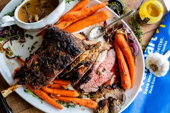 Roast Leg of Lamb with Blackened Honey Dijon Crust from The Food Charlatan. #ad #OhMaille #MyMaille #IDidItMailleWay Roast leg of lamb makes an unforgettable main dish for your Easter celebration! Roast lamb is so delicious, moist, and tender when it is done right. The secret is to turn the oven to 500 degrees to get that blackened honey dijon crust made with Maille® Honey Dijon, then lower it to get the most tender, juicy roast lamb you've ever had. #Walmart #leg #recipes #dinner #easter #lamb roast lamb on a platter with cooked carrots, herbs, gravy, and mustard.