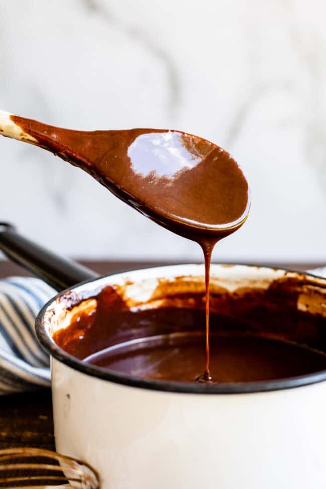Homemade Chocolate Sauce (The Only Hot Fudge Sauce You'll Ever Need) from The Food Charlatan. Once you go homemade, there's NO going back! This recipe is super easy, uses pantry ingredients, and takes 15 minutes. It's 10x better than store bought hot fudge sauce. It's creamy, smooth, and so chocolatey. #hotfudge #fudge #hot #icecream #Toppings #sauce #homemade #recipe #chocolate #Chocolatesauce #easy #desserts #sundae #uses #oldfashioned #withevaporatedmilk #best #topping #nocornsyrup homemade chocolate sauce drizzling off a spoon into a pot.