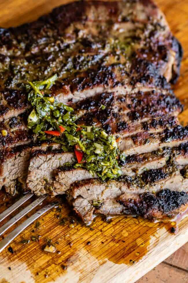 How to Cook Flank Steak (Grilled or Oven Broiled) from The Food Charlatan. Flank Steak is incredibly tasty when done well, but can be really easy to mess up. I will show you all my tips to get a flavorful, juicy, and tender flank steak, grilled outside or under the broiler! Using a great marinade, slicing correctly, and cooking hot and fast are the secrets to a fabulous flank steak. #flank #flanksteak #steak #beef #grill #grilled #easy #recipe #summer #marinade #oven #howtocook #chimichurri grilled sliced flank steak on a wooden cutting board with a metal fork, topped with chimichurri sauce.