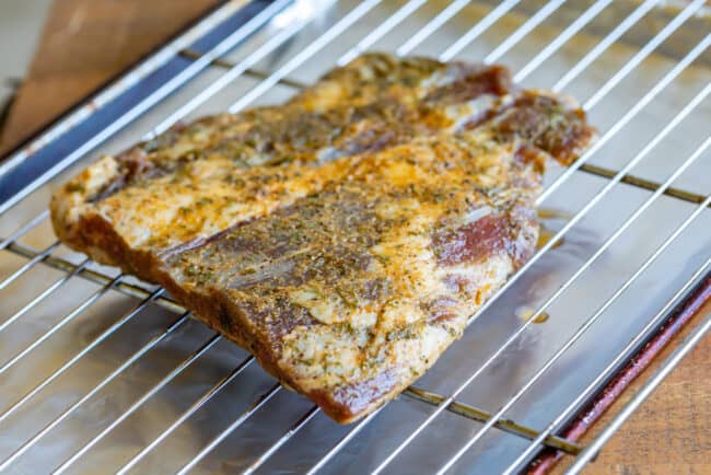 How to Cook Flank Steak (Grilled or Oven Broiled) from The Food Charlatan. Flank Steak is incredibly tasty when done well, but can be really easy to mess up. I will show you all my tips to get a flavorful, juicy, and tender flank steak, grilled outside or under the broiler! Using a great marinade, slicing correctly, and cooking hot and fast are the secrets to a fabulous flank steak. #flank #flanksteak #steak #beef #grill #grilled #easy #recipe #summer #marinade #oven #howtocook #chimichurri a marinated flank steak on a cooling rack over a baking sheet, ready to be broiled in the oven.