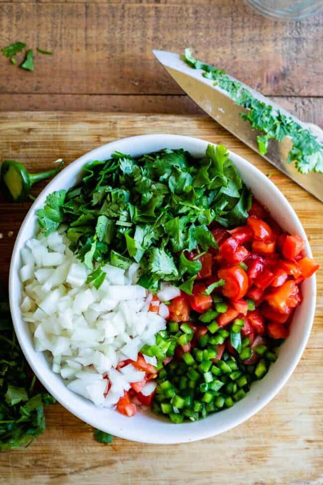 How to Make Authentic Pico de Gallo from The Food Charlatan. Learn how to make the best Pico de Gallo! It's a very simple Mexican salsa with tomatoes, onions, jalapeno, cilantro, and lime juice. Humble ingredients come together to make one of the best appetizers of all time! You will never look at tacos the same after topping them with this magical stuff. #picodegallo #salsa #Mexican #cold #fresh #appetizer #dip #forparties #easy #recipe #receta #authentic #best #homemade #restaurant #picodegalo pico de gallo ingredients in a white bowl with a wooden cutting board and knife.