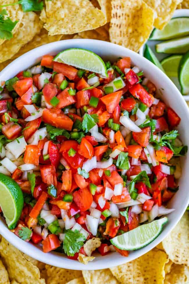 How to Make Authentic Pico de Gallo from The Food Charlatan. Learn how to make the best Pico de Gallo! It's a very simple Mexican salsa with tomatoes, onions, jalapeno, cilantro, and lime juice. Humble ingredients come together to make one of the best appetizers of all time! You will never look at tacos the same after topping them with this magical stuff. #picodegallo #salsa #Mexican #cold #fresh #appetizer #dip #forparties #easy #recipe #receta #authentic #best #homemade #restaurant #picodegalo pico de gallo in a white bowl with lime wedges and tortilla chips.