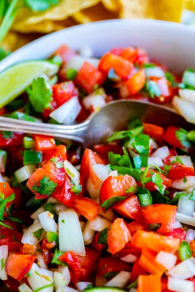 How to Make Authentic Pico de Gallo from The Food Charlatan. Learn how to make the best Pico de Gallo! It's a very simple Mexican salsa with tomatoes, onions, jalapeno, cilantro, and lime juice. Humble ingredients come together to make one of the best appetizers of all time! You will never look at tacos the same after topping them with this magical stuff. #picodegallo #salsa #Mexican #cold #fresh #appetizer #dip #forparties #easy #recipe #receta #authentic #best #homemade #restaurant #picodegalo pico de gallo in a white bowl with a metal spoon.