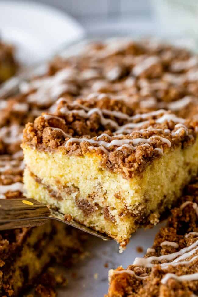 Sour Cream Coffee Cake, with a Ridiculous Amount of Streusel from The Food Charlatan. This is my FAVORITE recipe for Sour Cream Coffee Cake! My main complaint with Coffee Cake is that there is usually too much cake, not enough streusel. This recipe gives you the max amount of streusel without ruining the light fluffiness of the cake! A vanilla drizzle finishes it off! #coffeecake #sourcream #icing #frosting #drizzle #easy #recipes #cinnamon #streusel #sourcream #crumb #crumble #moist #breakfast Streusel topped coffee cake slice being lifted on a spatula from pan.