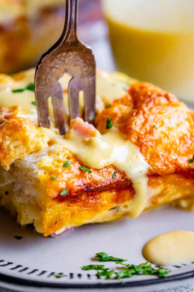 Cheesy Ham and Egg Breakfast Casserole with Biscuits from The Food Charlatan. This crowd-pleasing casserole is my new favorite! Use store bought or homemade biscuits to make a moist and delicious brunch. Top it all off with Hollandaise Sauce for an amazing holiday breakfast! #breakfast #brunch #ham #bacon #sausage #easy #recipe #holidays #Thanksgiving #Christmas #makeahead #withbiscuits #overnight #recipes #foracrowd #best #nightbefore #simple #quick #forlargegroups #Pillsbury #freezer #savory biscuit, ham, and egg breakfast casserole with a fork in the top and hollandaise melting off.