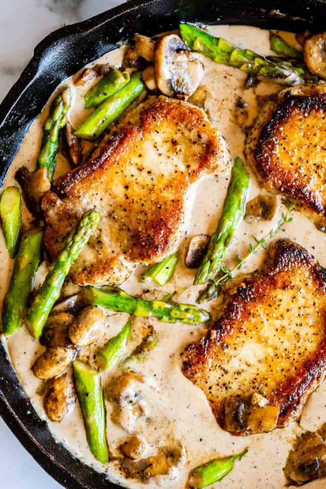 Creamy Pan Seared Pork Chops with Asparagus from The Food Charlatan. Creamy Pan Seared Pork Chops is such an easy weeknight dinner! Juicy boneless pork chops are seared over high heat to get that irresistible crust. Sautéed mushrooms and asparagus in a creamy pan sauce bring it all together. Dinner is done in about 30 minutes! #panseared #porkchop #easy #recipe #boneless #healthy #skillet #balsamic #creamy #asparagus #mushroom #pansauce #onthestove #stovetop #cream #pepper #garlic #weeknight seared boneless pork chops with asparagus, mushrooms, fresh thyme, and creamy gravy in a cast iron skillet.