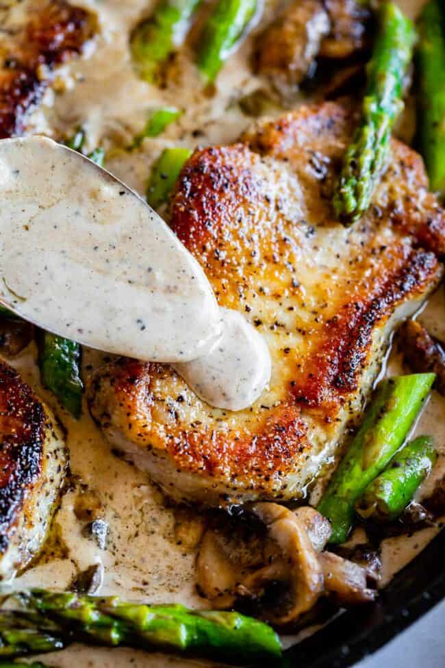 Creamy Pan Seared Pork Chops with Asparagus from The Food Charlatan. Creamy Pan Seared Pork Chops is such an easy weeknight dinner! Juicy boneless pork chops are seared over high heat to get that irresistible crust. Sautéed mushrooms and asparagus in a creamy pan sauce bring it all together. Dinner is done in about 30 minutes! #panseared #porkchop #easy #recipe #boneless #healthy #skillet #balsamic #creamy #asparagus #mushroom #pansauce #onthestove #stovetop #cream #pepper #garlic #weeknight seared pork chops with asparagus, mushrooms, and a creamy gravy being spooned on top.
