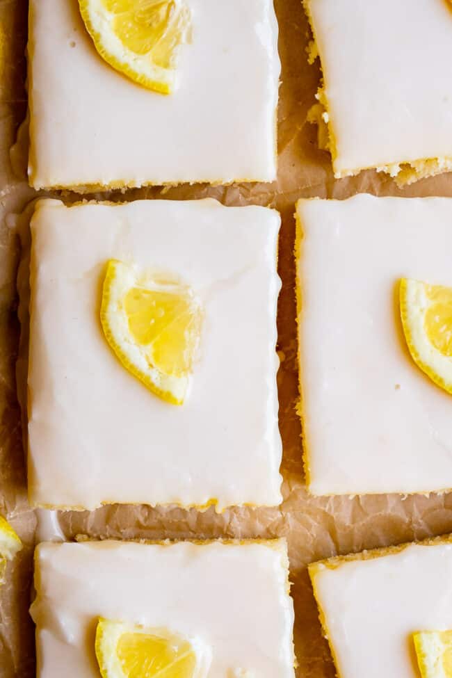 Lemon Sheet Cake with Lemon Glaze from The Food Charlatan. This Lemon Sheet Cake with Lemon Glaze is perfect for lemon lovers! A supremely lemony and MOIST cake, paired with a bright lemon glaze. It is perfect for summertime, or anytime! This recipe feeds a crowd and is super easy to make. #lemon #cake #sheetcake #lemoncake #citrus #summer #easy #recipe #zest #tart #withglaze #fromscratch #homemade #glaze #best #Texas #simple #dessert #feedsacrowd #foracrowd squares of lemon sheet cake, glazed, with lemon slices.