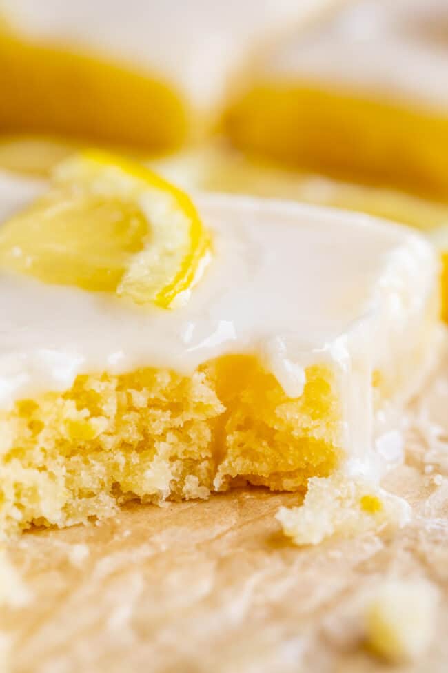 Lemon Sheet Cake with Lemon Glaze from The Food Charlatan. This Lemon Sheet Cake with Lemon Glaze is perfect for lemon lovers! A supremely lemony and MOIST cake, paired with a bright lemon glaze. It is perfect for summertime, or anytime! This recipe feeds a crowd and is super easy to make. #lemon #cake #sheetcake #lemoncake #citrus #summer #easy #recipe #zest #tart #withglaze #fromscratch #homemade #glaze #best #Texas #simple #dessert #feedsacrowd #foracrowd a piece of lemon sheet cake with glaze and a lemon slice with a bite taken out.