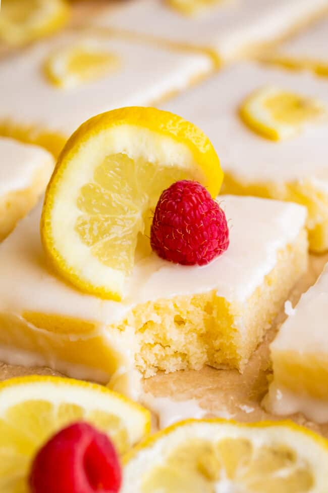 Lemon Sheet Cake with Lemon Glaze from The Food Charlatan. This Lemon Sheet Cake with Lemon Glaze is perfect for lemon lovers! A supremely lemony and MOIST cake, paired with a bright lemon glaze. It is perfect for summertime, or anytime! This recipe feeds a crowd and is super easy to make. #lemon #cake #sheetcake #lemoncake #citrus #summer #easy #recipe #zest #tart #withglaze #fromscratch #homemade #glaze #best #Texas #simple #dessert #feedsacrowd #foracrowd squares of glazed lemon sheet cake with lemon slices and fresh raspberries, a bite missing from one piece.
