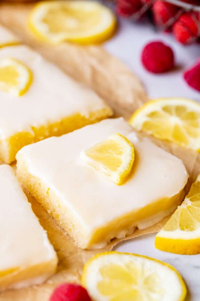 Lemon Sheet Cake with Lemon Glaze from The Food Charlatan. This Lemon Sheet Cake with Lemon Glaze is perfect for lemon lovers! A supremely lemony and MOIST cake, paired with a bright lemon glaze. It is perfect for summertime, or anytime! This recipe feeds a crowd and is super easy to make. #lemon #cake #sheetcake #lemoncake #citrus #summer #easy #recipe #zest #tart #withglaze #fromscratch #homemade #glaze #best #Texas #simple #dessert #feedsacrowd #foracrowd squares of lemon sheet cake with lemon slices and fresh raspberries on a parchment lined baking sheet.