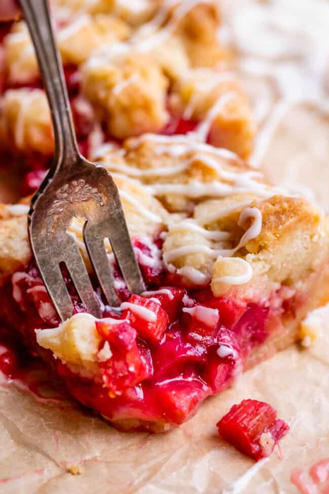Rhubarb Shortbread Bars from The Food Charlatan. Rhubarb Shortbread Bars are a dream come true! A thick and buttery shortbread crust, topped with a layer of simple rhubarb filling, and topped with even more glorious shortbread. There is a secret ingredient in this recipe that makes these rhubarb bars bright ruby red, and it's not food coloring. It works like a charm! #easy #recipe #rhubarb #shortbread #butter #bars #dessert #withoutstrawberry #recipe #shortbreadcrust #lemon #best #pie fork slicing into rhubarb crumble bars with shortbread pastry