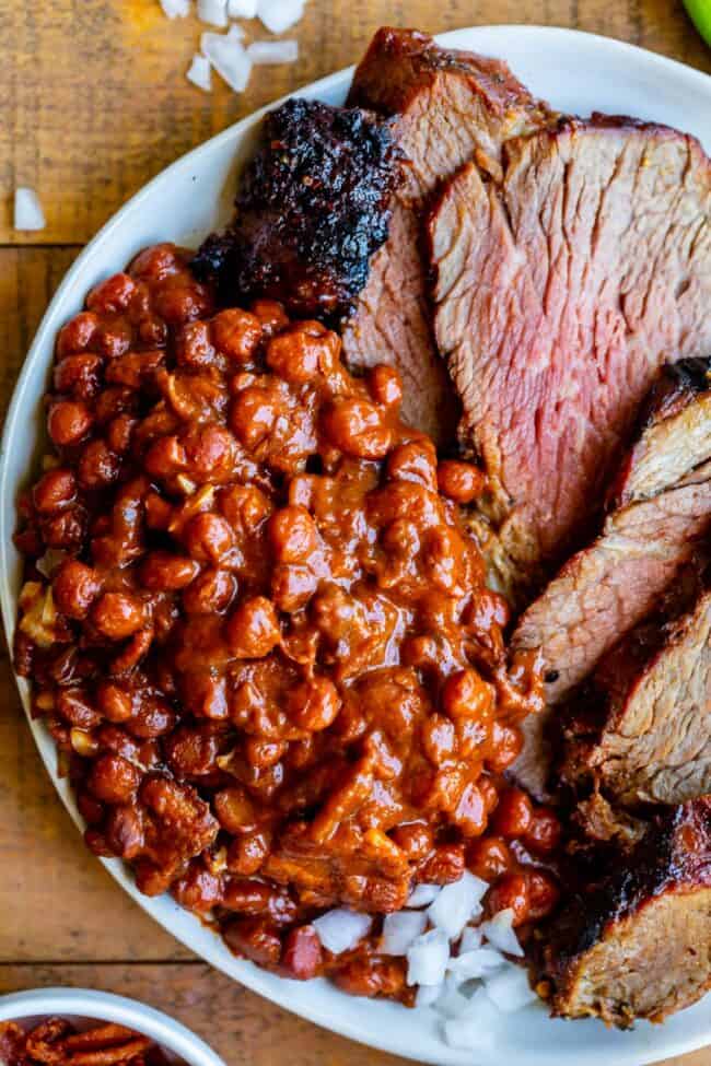 Santa Maria Pinquito Beans from The Food Charlatan. These Santa Maria style Pinquito Beans are the perfect summer barbecue side dish! Originally created in Santa Maria, California to be served alongside Grilled Tri Tip, these beans are smokey and sweet with the perfect blend of spices. Eat them with tri tip or serve alongside whatever else you're barbecuing this summer! #easy #recipe #bbq #barbecue #beans #bakedbeans #santamaria #tritip #sidedish #bacon #pinquito #creamy #summer #onion #anaheim santa maria beans on a plate with tri tip.