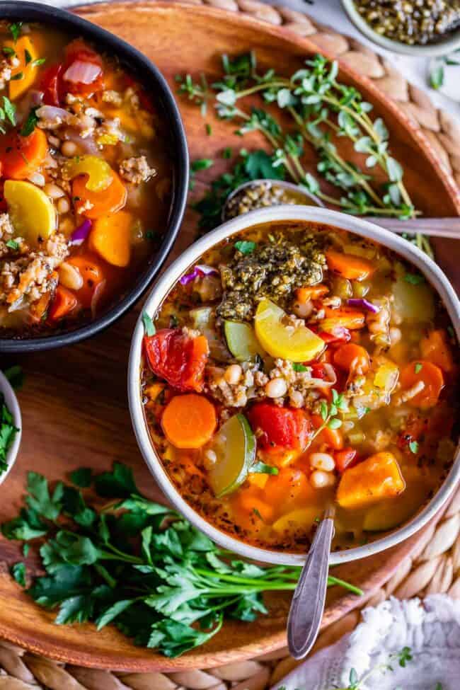 ground turkey vegetable soup in two bowls on a wooden charger plate with fresh herbs on the side.